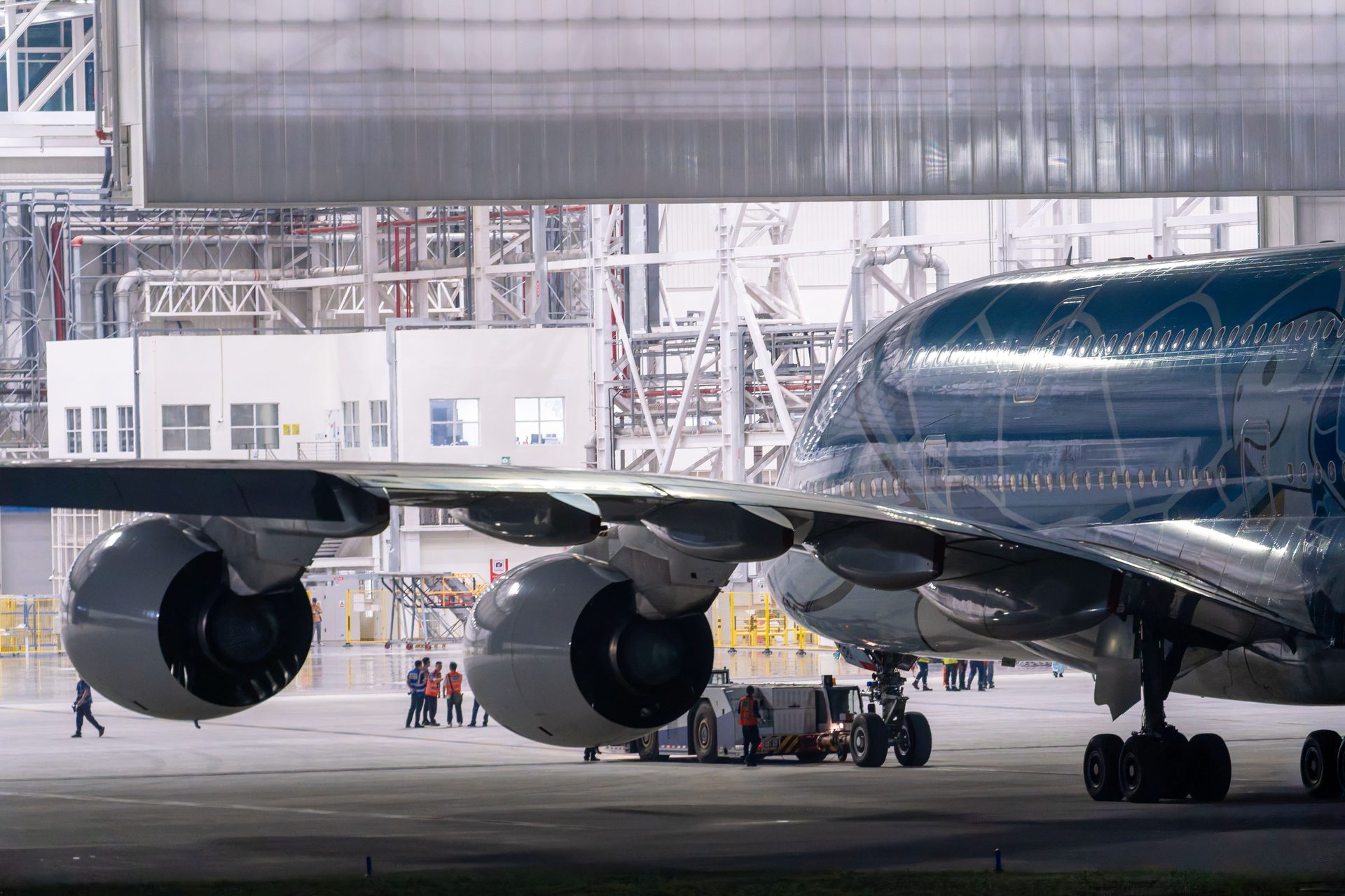 Wide-body jet parked inside a bright maintenance hangar with ground crew at work